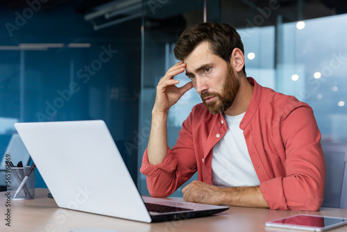 Obraz na plátně A stressed and tired young businessman, student, freelancer in a red shirt sits in the office at a laptop