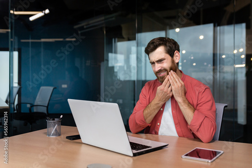 A young man, businessman, freelancer, designer sits in the office at the table. works on a laptop. He holds his cheek, feels discomfort in his mouth, has a severe toothache.