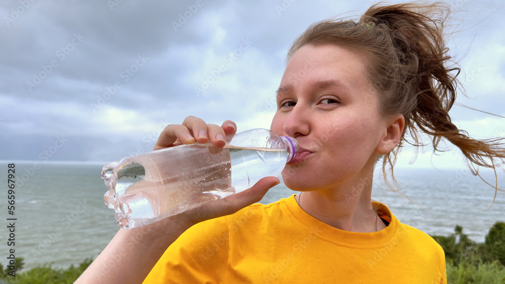 Portrait of happy positive thirsty girl, young beautiful woman is drinking pure fresh mineral water from plastic bottle on natural sea, ocean background in tropical country, holding bottle in hand Portrait of happy positive thirsty girl, young beautiful woman is drinking pure fresh mineral water from plastic bottle on natural sea, ocean background in tropical country, holding bottle in hand