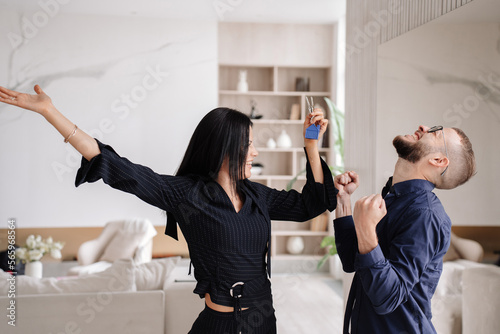 Young caucasian couple celebrating new apartment. Excited Italian woman holds keys of flat dancing looks at husband. Handsome beardy man clenches fists like winner after purchasing home. Mortgage