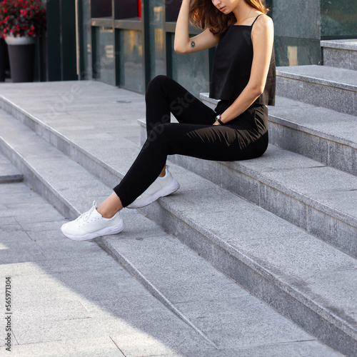 Young stylish happy brunette woman sitting on a busy business urban street in black sleeveless top and pants. Street style photoshoot. © nickitavanat