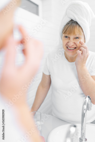Gorgeous mid age older adult 50 years old blonde woman wears bathrobe in bathroom applying nourishing antiage face skin care cream treatment  looking at mirror doing daily morning beauty routine.