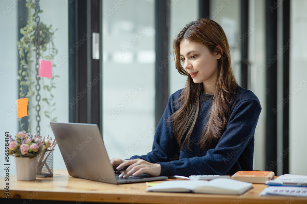 © SOMKID - Happy smiling young asian business woman using laptop sitting at desk in the office, Company worker woman.