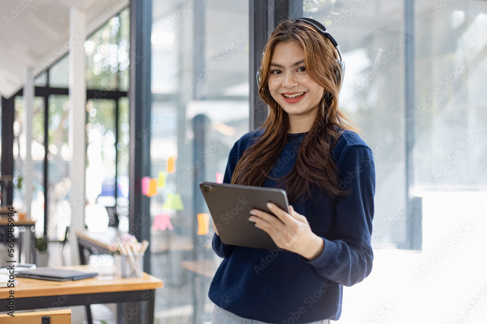 Fototapeta premium Young woman smiling and holding digital tablet with earphones standing in office, People And Technology Concepts. 