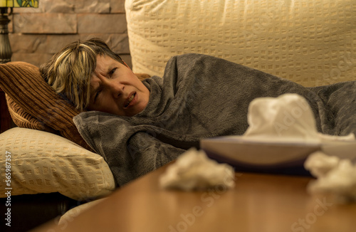 mujer mediana edad enferma con dolores  en el sofa de casa tapada con una manta, tomando medicina mirando el movil y el ordenador photo
