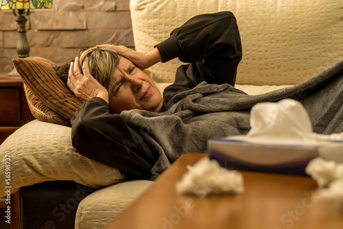 mujer mediana edad enferma con dolores  en el sofa de casa tapada con una manta, tomando medicina mirando el movil y el ordenador photo