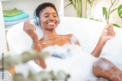 Fotografia Attractive black woman wearing headphones happy relaxing and dancing in foam bath in beautiful bathroom with plants