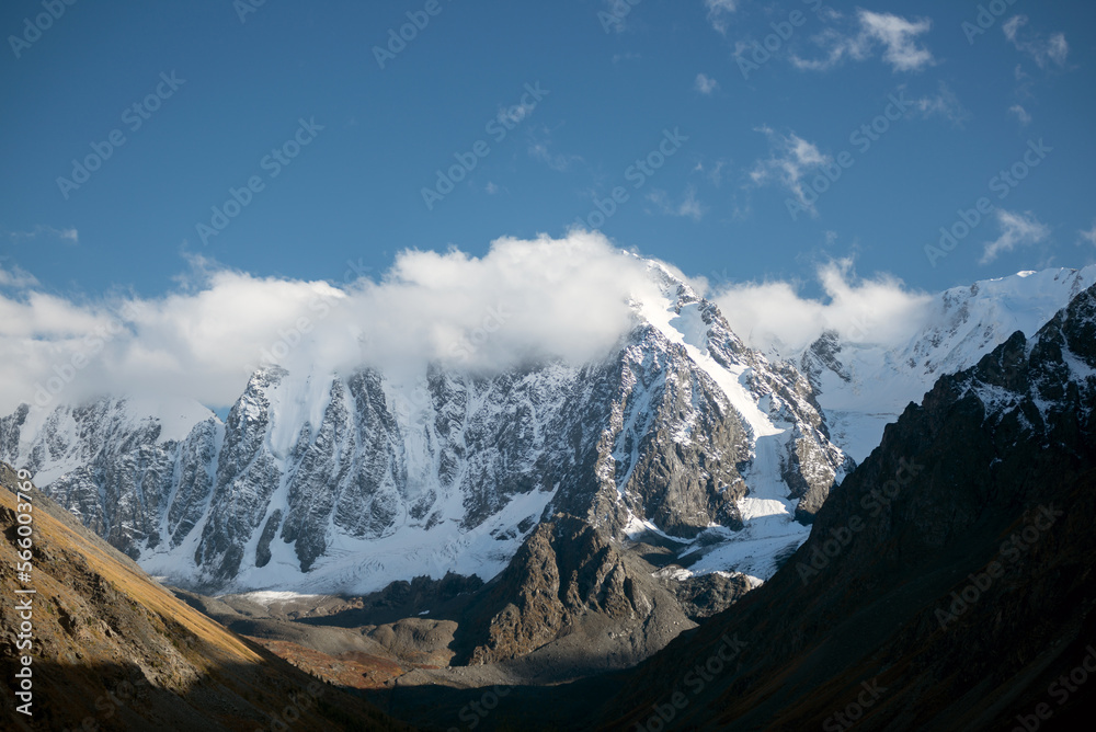 Panorama white clouds cover the tops of high rocky mountains with snow and glaciers in Altai in autumn in the shade during the day. Peaks Dream, Fairy Tale, Beauty.
