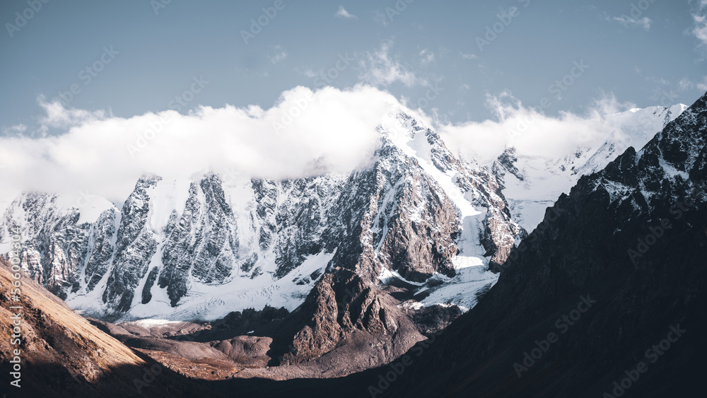 White clouds cover the tops of high rocky mountains with snow and glaciers in Altai with fog.