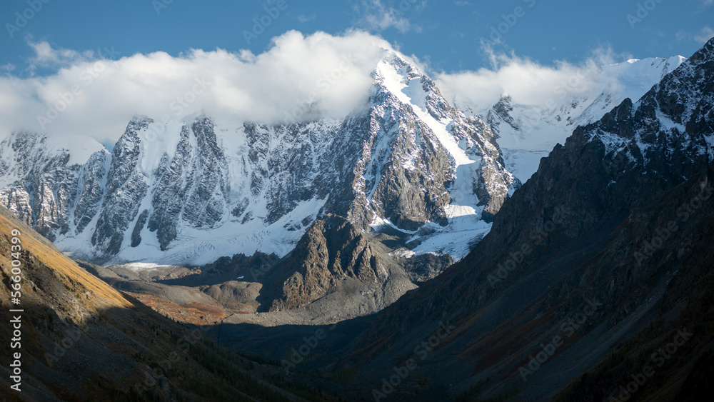 White clouds cover the peaks of high rocky mountains with snow and tongue of glaciers in the Altai in the shade.