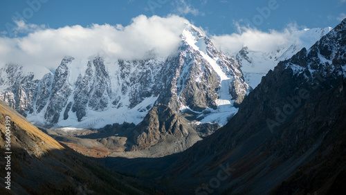 White clouds cover the peaks of high rocky mountains with snow and tongue of glaciers in the Altai in the shade.