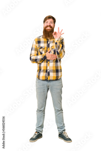 Young adult redhead man with a long beard standing full body isolated winks an eye and holds an okay gesture with hand.