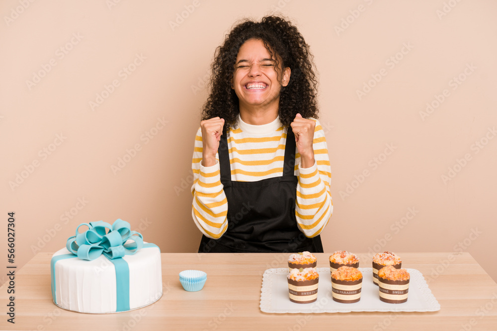 Young african american woman preparing a sweet cake and muffins on a table celebrating a victory, passion and enthusiasm, happy expression. Young african american woman preparing a sweet cake and muffins on a table celebrating a victory, passion and enthusiasm, happy expression.