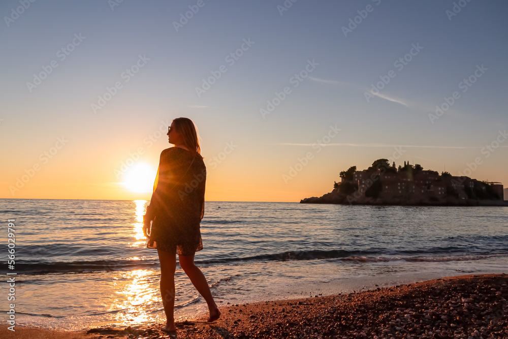Silhouette of barefoot woman on sand beach with scenic view on idyllic island Sveti Stefan at romantic sunset, Budva Riviera, Adriatic Mediterranean Sea, Montenegro, Europe. Summer vacation at seaside