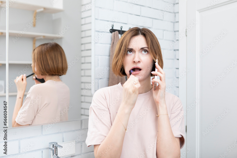 Young attractive woman talking on phone while brushing teeth in the bathroom. Morning routine concept Young attractive woman talking on phone while brushing teeth in the bathroom. Morning routine concept