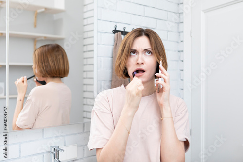 Young attractive woman talking on phone while brushing teeth in the bathroom. Morning routine concept