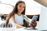 Smiling woman freelancer holding mobile phone using laptop working online sitting at workplace. Portrait of Young happy designer looking at camera, successful business