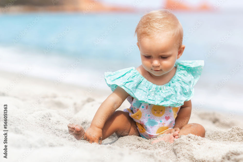 Beautiful baby girl playing with sand while sitting in a swimsuit on the beach in summer. Summer weekend, family travel, vacation concept Beautiful baby girl playing with sand while sitting in a swimsuit on the beach in summer. Summer weekend, family travel, vacation concept