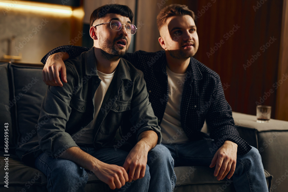 Two men are sitting on the couch at home and watching TV. A great TV show, the guys are emotionally tense and intriguingly looking at the screen. Two men are sitting on the couch at home and watching TV. A great TV show, the guys are emotionally tense and intriguingly looking at the screen.