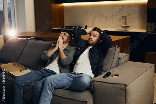 Фотография Two men are sitting on the sofa at home and watching TV and eating pizza