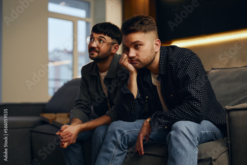 Two men are sitting on the sofa at home and watching TV and eating pizza. The emotion of shock, sadness, disappointment.