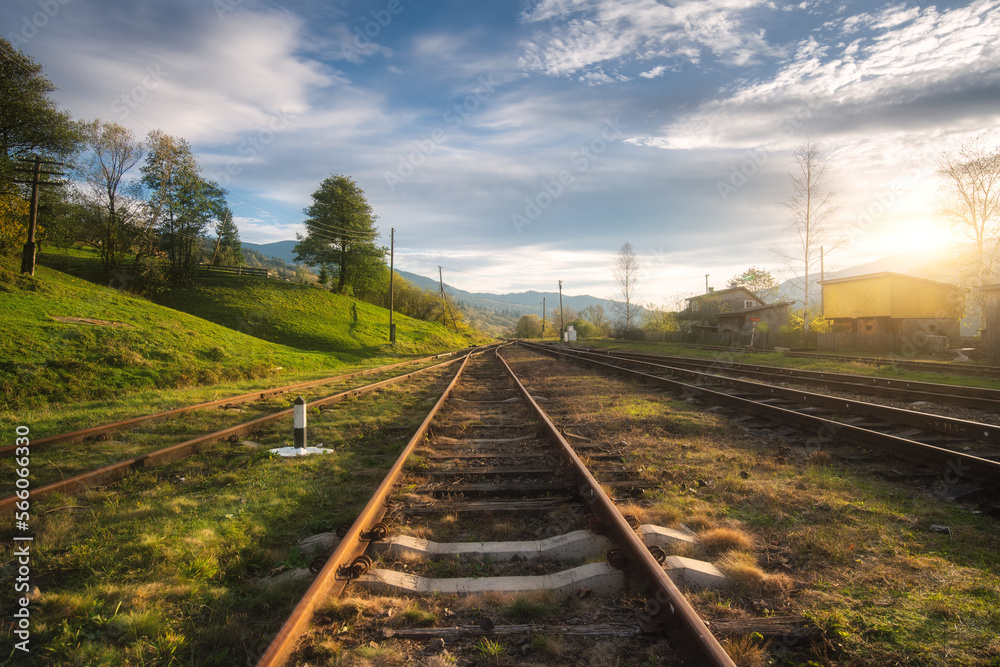 Railroad in mountains at sunset in summer. Beautiful industrial landscape with railway station, trees, green grass, blue sky, clouds in spring. Old rural railway platform in Ukraine. Transportation