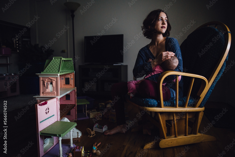 Breastfeeding mom sitting in rocking chair near little girl dollhouse