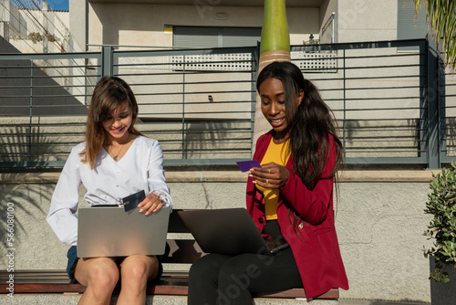 Two smiling businesswomen sitting on a bench outdoor shop online with laptop and credit card.