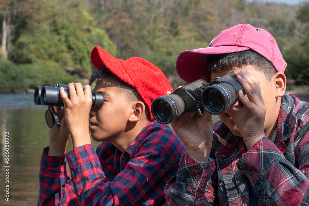 Obraz premium Asian teen boys hold the national park map stand by sling bridge, reading details of birdwatching on map before using their binoculars to watch the bird and fish, summer vacation and trekking concept.
