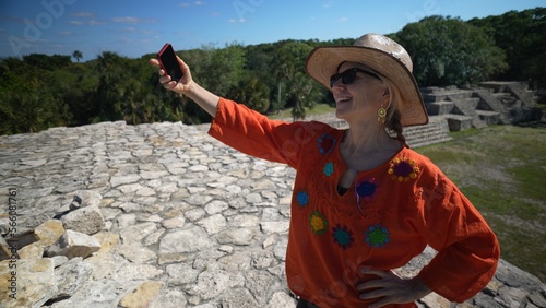 Pretty mature woman wearing ethnic clothes, sunglasses, hat taking selfie at the top of Xcambo Mayan pyramid ruins. Adventure is ageless. photo