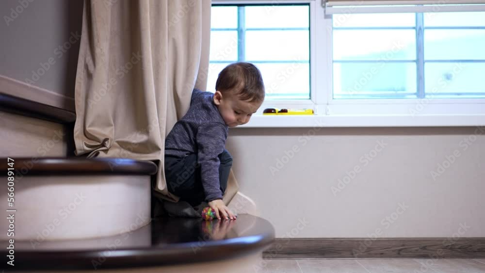 Caucasian toddler boy stands on the stair near the window. Child takes the rubber ball and carefully descends on the floor. Caucasian toddler boy stands on the stair near the window. Child takes the rubber ball and carefully descends on the floor.