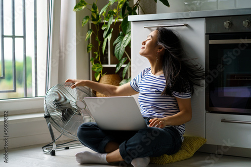Satisfied Asian woman sits on floor with laptop on crossed knees, enjoying wind from electric fan. Relaxed Korean female freelancer takes break from work, enjoys the fresh breeze from fan. Summertime