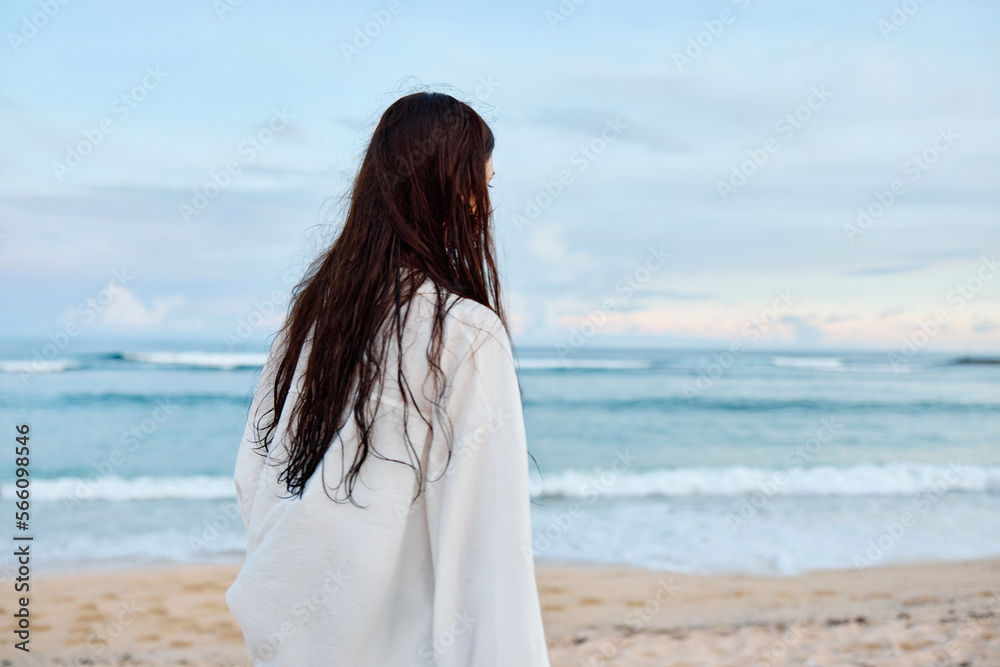 Happy tanned slender woman in white swimsuit shirt and denim shorts walks on the beach with her back to the camera on the sand by the ocean with wet hair after swimming, sunset light Happy tanned slender woman in white swimsuit shirt and denim shorts walks on the beach with her back to the camera on the sand by the ocean with wet hair after swimming, sunset light