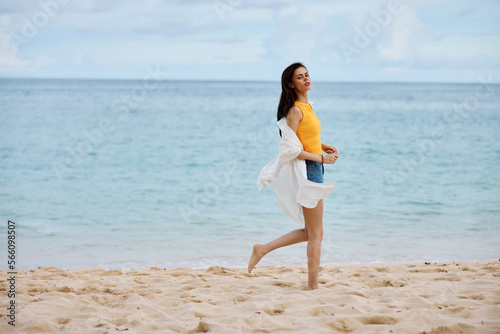 Sports woman runs along the beach in summer clothes on the sand in a yellow tank top and denim shorts white shirt flying hair ocean view  beach vacation and travel