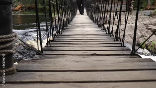 Man walking on suspension bridge made of wooden slats over waterfall. Male legs crossing stormy river along swinging hanging bridge. Human moving straight towards the camera. photo