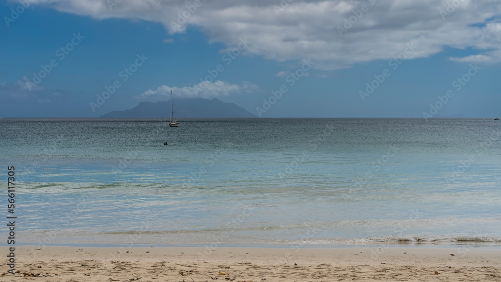 A yacht is visible on the surface of the calm turquoise ocean. The outline of the island on the horizon. Foam of waves on the sand of the beach. Clouds in the blue sky. Seychelles. Mahe. Beau Vallon