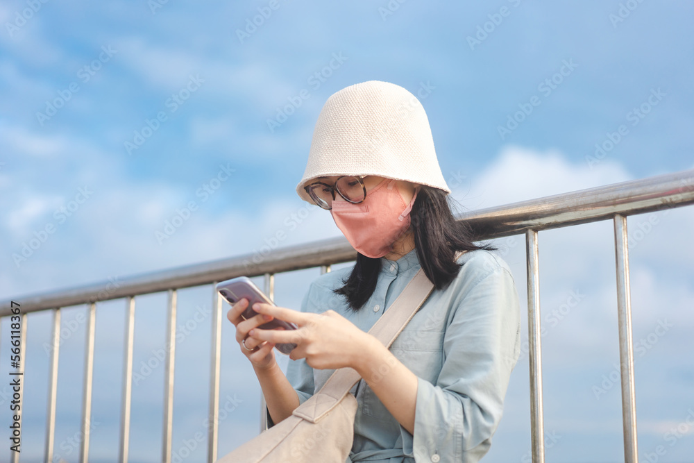 Asian woman wearing face mask using smartphone at outdoor with blue sky background