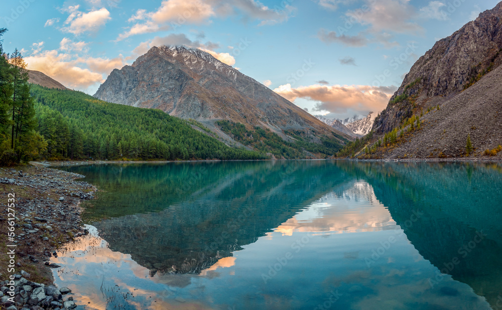 Panorama of the turquoise transparent lake Shavlinskoe in the shade with stones among the mountains with reflection of the peaks with glaciers and snow in Altai.
