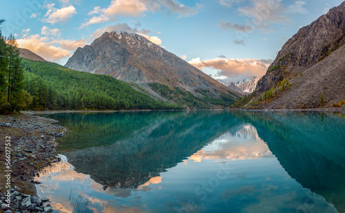 Panorama of the turquoise transparent lake Shavlinskoe in the shade with stones among the mountains with reflection of the peaks with glaciers and snow in Altai.