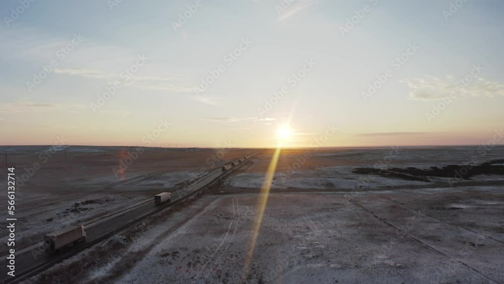 A convoy of trucks carrying goods is passing at dawn into the distance along the road in the steppe covered with snow illuminated by the rays of the sun, side view, rear A convoy of trucks carrying goods is passing at dawn into the distance along the road in the steppe covered with snow illuminated by the rays of the sun, side view, rear