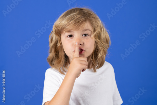 Kid with show no talk gesture. Boy with shows shh sign. Be quiet. Hush dont tell. Child put finger to lips mouth, ask stop share rumor, isolated on blue studio isolated background.
