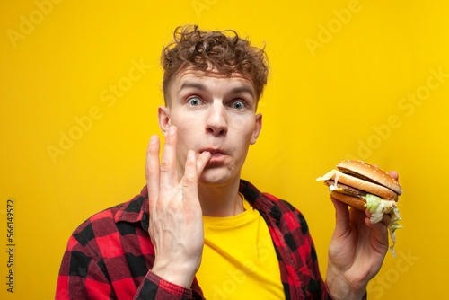 Fotografie surprised guy student eats a big tasty burger on a yellow background and licks t