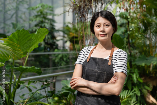 half length portrait of a confident asian Japanese female gardener wearing apron and looking at camera with folded arms at background of tropical greenhouse
