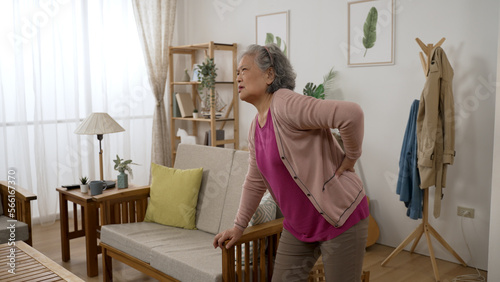 asian senior lady is feeling pain for she sprained her lower back while she is walking into the living room at home and supporting herself with sofa armrest