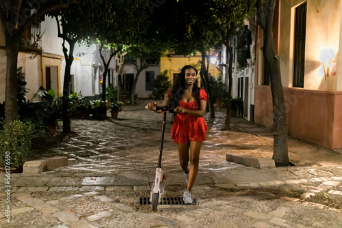 Full length of cheerful girl wearing in red dress walking with electric scooter enjoying summer night outdoor in old downtown street photo