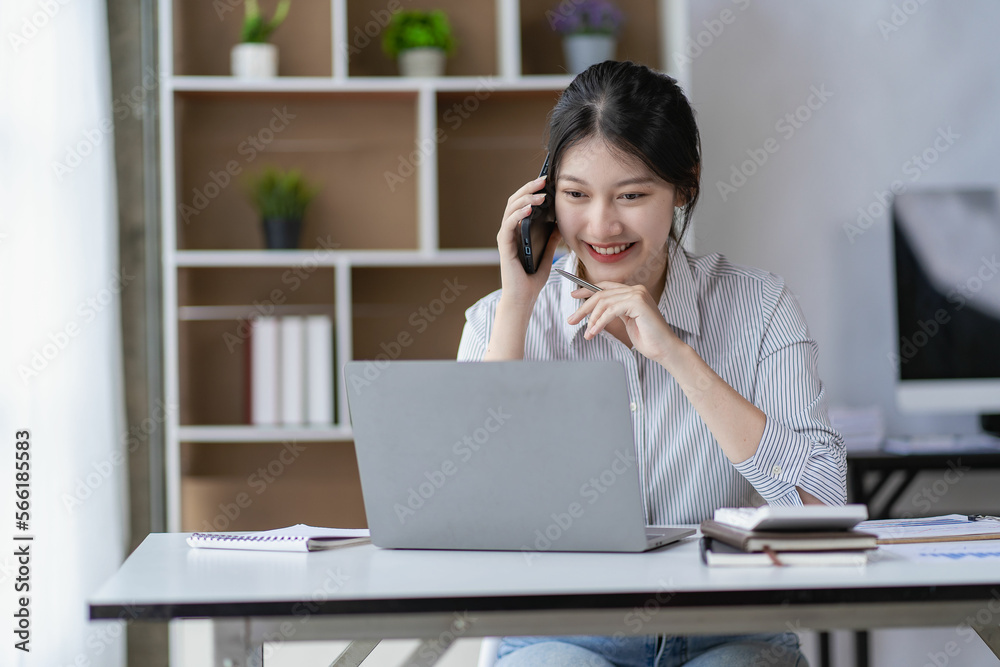 Asian woman with black hair holding a smart phone Phone talking to work team with laptop and financial graph paperwork in analyzing and calculating at desk. Asian woman with black hair holding a smart phone Phone talking to work team with laptop and financial graph paperwork in analyzing and calculating at desk.