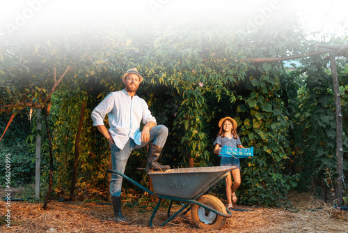 Web banner of organic gardening. Handsome bearded gardener poses with his foot on a cart. Little girl comes from geenhouse with harvest box. The concept of summer and harvesting