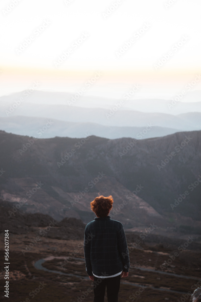 Young redhead man wearing a dark flannel looking at the sunset view from the top of a mountain with mountain layers in the background in Las Villuercas, Cáceres, Extremadura, Spain