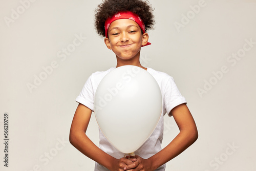 Portrait of funny sporty african boy kid making faces holding white helium balloon with copy space in hands blowing cheeks, trying no to laugh isolated on gray studio background © Anatoliy Karlyuk