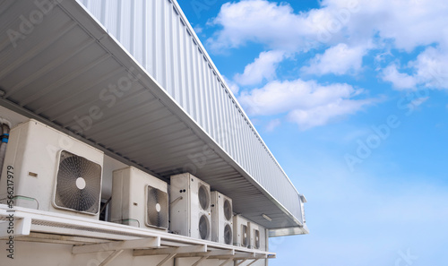 Row of air conditioner compressors on white wall under steel roof eaves of modern convenience store against blue sky in perspective side view